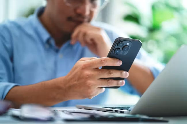 A person in a blue shirt is holding a smartphone in one hand while resting their chin on the other hand, appearing focused on the device. In the background, there is a laptop and greenery.